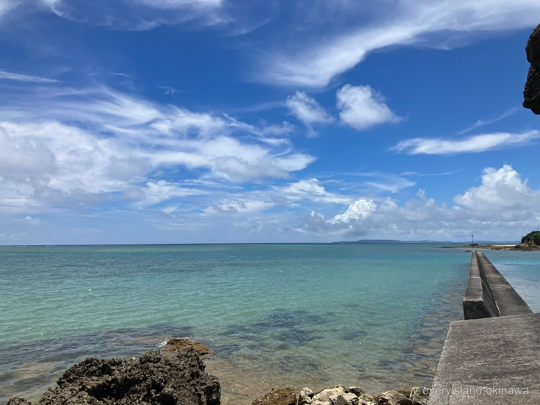Emerald shallows and a low breakwater on the Ginoza coast under summer clouds